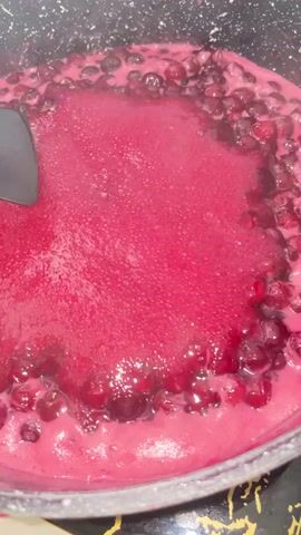 This is a close-up video of a dark pot with a bubbling, deep pink mixture, likely jam or compote being made. Dark red berries, possibly cranberries or cherries, are visible throughout the mixture, particularly around the edges of the pot. A black spatula is used to stir the contents. The setting appears to be a kitchen, with a black and gold patterned surface visible at the bottom of the frame. The lighting suggests it could be daytime or indoor lighting. No people are visible. There is no visible text in the video. The activity is the cooking and reduction of a berry mixture.