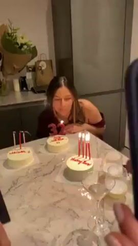 A woman in a maroon dress is celebrating her 32nd birthday in a kitchen. Three small, round cakes are on the marble countertop in front of her. Each cake has white frosting and red writing. The cakes are labeled "Adriam," "32," and "Happy Birthday," respectively. Each cake also has candles that have just been blown out and are smoking. To the side of the cakes are several glasses, a martini glass, and a vase of flowers. Behind the woman are kitchen cabinets and a refrigerator. Another person is partially visible in the foreground, holding a phone to record the scene. The woman claps her hands and smiles.