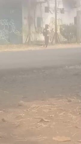 The video shows a dusty street scene. In the foreground, there is dry dirt with scattered leaves and twigs. In the middle ground, a paved road is visible, also covered in dust. A person is seen walking on the side of the road. In the background, there is a dilapidated building with graffiti on its walls. A truck is seen driving on the road. The lighting suggests it is daytime. The overall atmosphere is hazy, likely due to dust or smoke.