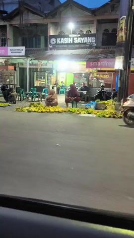 A video captures a street scene at dusk. In the foreground, a road is visible. In the background, a row of shops lines the street. Several people are seated on the ground on blue tarps, surrounded by large piles of bananas. Two individuals are wearing headscarves and appear to be selling the fruit. Behind them, a motorcycle is parked. The shops display various signs, including "smartfren," "RUMAH MAKAN MUHAMMAD," and "WARKOP KASIH SAYANG." Bright lights illuminate the area, suggesting the shops are open. The overall atmosphere is that of a bustling night market or roadside vendor scene.
