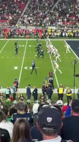 The video shows a football game from the perspective of someone in the stands. The field is green with white yard lines and numbers. Players in dark blue and light colors are lined up on the field. Some run, others stand still. There are officials wearing black and white stripes. There are people standing near the field, in dark suits or with yellow jackets. The stands are full of spectators. The perspective is from a higher level, giving a wide view of the field. The sky is not visible.