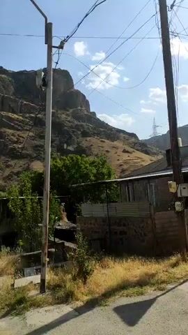 The video shows a rural scene with mountains in the background. A power line pole stands in the foreground, with wires extending from it. There are some buildings and trees visible, as well as a road. The sky is clear and blue. In one shot, two people are walking down the road in the distance.