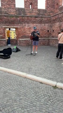 The video captures a street performer playing an accordion and guitar in front of a weathered brick wall adorned with battlements. The cobblestone ground suggests an outdoor public space, possibly a historical site given the architecture. A guitar case and an amplifier are placed on the ground near the performer.

Several people are present, observing the performance or passing by. One person stands in a rectangular opening in the wall, looking out. Others are seated on a ledge along the wall or walking in the background. The lighting indicates it is daytime, likely late afternoon or early evening, with a soft glow. The overall atmosphere appears relaxed and leisurely, with the music adding to the ambiance of the setting. The specific location is hinted at as Verona, Italy, which aligns with the architectural style. No visible text is present in the video.