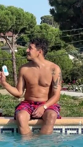 A man with tattoos on his left arm sits shirtless on the edge of a swimming pool, his legs submerged in the water. He holds a small, white object with blue accents in his right hand. To his left, another person, partially visible, also sits on the pool edge with their legs in the water. Bubbles rise from the water between the two individuals. The environment appears to be outdoors, with lush green trees and houses visible in the background under a clear sky. The man is looking to his left, away from the camera.