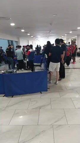 This is a first-person perspective video of a crowded indoor area, possibly a mall or convention center. The camera operator is walking through the space, which has a tiled floor and a long ceiling with recessed lighting. Several people are visible, some standing in groups, others walking. In the foreground, a blue barrier with a white logo is set up, with people standing on the other side. Some individuals are wearing casual clothing like t-shirts and shorts, while others are in more formal attire. There are tables and chairs set up in the background, and some people appear to be interacting with each other or with displays. The overall atmosphere seems to be one of a public event or gathering.