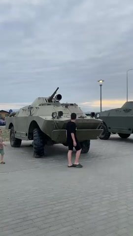 A person walks past two military vehicles. The vehicle in the foreground is a light green armored personnel carrier with a mounted gun. The vehicle behind it is also military, and a third, similar vehicle is partially visible to the right. The ground is paved with light gray bricks. The sky is cloudy with some yellow light visible on the horizon, suggesting dawn or dusk. The person is wearing a black t-shirt and shorts.