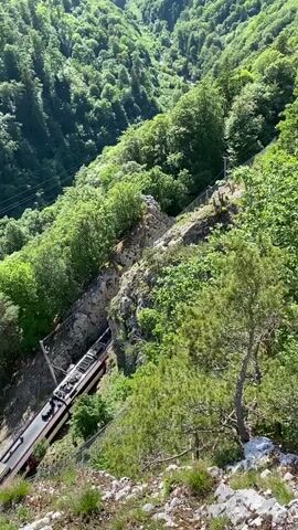 The video shows a steep, forested valley. A train passes along a track cut into the mountainside. The view is from above the train, looking down at it and across the valley to the opposite, equally steep, forested slope. The sky is blue with a few wispy clouds. The train is long and light-colored with red and black stripes. A fence runs along the edge of the track. There are a few people standing on the track near the train. The valley is very green and lush. The video tilts up to show more of the valley.