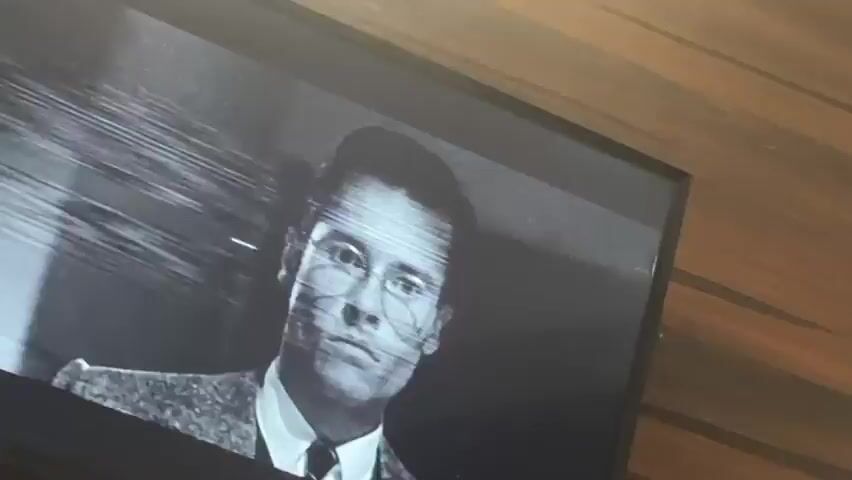 The video shows a black and white clip on a television screen. A man in a suit and hat is visible, speaking with intensity. Another individual stands beside him. Both are in what appears to be an outdoor setting, possibly a street. The video quality is compromised by horizontal lines across the screen. The backdrop is a wall with a wood-like texture.