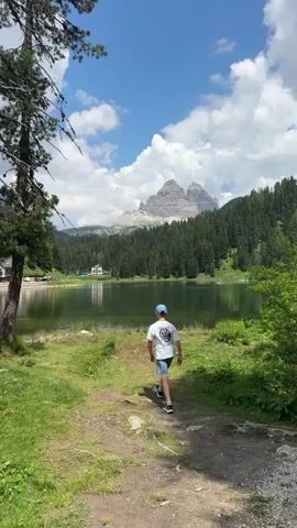This vertical, eye-level video shows a young boy walking away from the camera on a dirt path towards a serene lake. The boy is wearing a white t-shirt, grey shorts, a blue baseball cap, and dark sneakers. He walks with his hands in his pockets, suggesting a relaxed and casual demeanor.

The scene is set in a picturesque natural landscape. The lake, with its calm, reflective surface, occupies the midground. On the far side of the lake, a dense, dark green forest stretches across the rolling hills, creating a stark contrast against the bright blue sky. Beyond the forest, imposing, rugged mountains with rocky peaks rise dramatically. The mountains are partially obscured by fluffy white clouds, suggesting a pleasant day. A few buildings are visible on the far shore, nestled amongst the trees, hinting at human settlement in this otherwise wild environment.

The time of day appears to be daytime, likely late morning or early afternoon, given the bright sunlight and the position of the shadows. The weather is fair, with a clear blue sky punctuated by scattered white clouds. The lush green grass and foliage indicate a temperate season, likely summer. The overall mood is peaceful and idyllic, evoking a sense of natural beauty and tranquility.

There is no visible text in the media file. The location is identified as Auronzo di Cadore, Italy, a region known for its stunning alpine scenery, which is consistent with the visual elements in the video.