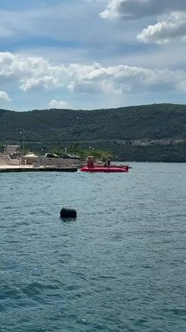The video shows a body of water with a black buoy in the foreground.  In the distance is a concrete pier with a building, some cars, and what appears to be a red boat or possibly a submarine.  There are people on the pier near the red boat.  Behind the pier is a hilly green landscape. The sky is blue with white clouds.