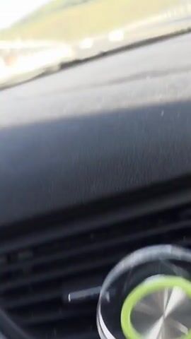 This video shows a view from inside a vehicle traveling on a highway. The camera is pointed forward, looking out through the windshield. The highway is paved with asphalt and has multiple lanes, separated by white lines. A metal guardrail runs along the side of the road. In the distance, other vehicles can be seen on the highway. The landscape on either side of the road consists of green hills, possibly farmland or vineyards, under a clear blue sky. The interior of the car, including the dashboard and speaker grille, is visible in the foreground. The video appears to be a first-person perspective of a road trip.