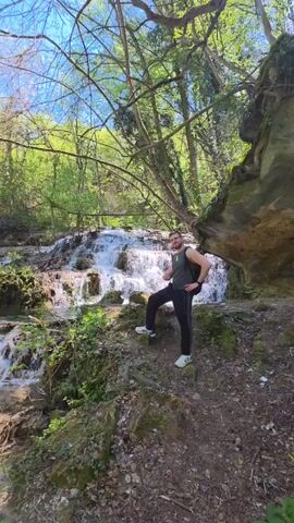 A man is standing in front of a waterfall in a forest. The man is wearing a gray tank top, black pants, and white shoes. He has a black backpack on his back. He is standing on a rocky outcrop. Behind the man, there is a waterfall cascading down a series of rocks. The water is white and foamy. The forest is lush and green. There are many trees and plants. The sky is blue and there are a few clouds.