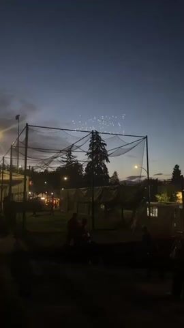 The video captures a twilight scene at an outdoor facility, possibly a sports field or entertainment venue. The sky above is a deep blue, transitioning into darker hues as night falls, with faint wisps of clouds visible on the left. In the sky, a formation of small, bright lights is arranged in a pattern that appears to be forming an image or text, though it is not entirely clear.

Dominating the foreground and midground is a large, black netting structure, likely a protective barrier for a sports field, such as a baseball or soccer field, or perhaps a batting cage. The netting is supported by a metal frame and extends upwards and outwards.

Beneath the netting, the ground is covered in grass, and several people are visible. Some are standing, while others appear to be seated or in motion. Their figures are largely silhouetted against the dim light, making specific identification difficult. They are casually dressed, suggesting a relaxed atmosphere.

In the background, there are trees, some of which are tall and prominent. There are also some structures, possibly buildings or tents, illuminated by artificial lights, indicating human activity and infrastructure. Streetlights are also present, casting pools of light on the ground and highlighting the edges of objects. The overall impression is one of an outdoor event or gathering taking place as evening sets in.