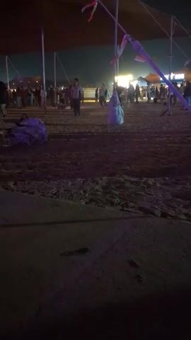A wide, low-angle shot shows a nighttime beach scene. Above, a large, dark awning is supported by metal poles with colorful ribbons. The ground is sand, with scattered debris. Several people are walking around and milling about. In the background, a crowd is gathered near illuminated structures. A person walks by the camera in the foreground. The lighting is dim, but highlights the sand and people in the middle ground.