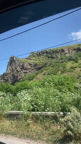 The video shows a view from inside a vehicle looking out at a rocky mountain landscape under a clear blue sky. Green trees and vegetation cover the slopes of the mountain and the foreground. The camera appears to be in motion, as indicated by the slight blur of the foreground vegetation.