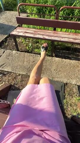 A first-person perspective video shows a person wearing a pink dress and black sandals sitting on a swing outdoors. Their legs are visible from the knees down. In front of them is a wooden bench with a reddish-brown frame, a worn wooden seat, and a similar backrest. The ground beneath the swing and bench is a mix of dirt, gravel, and scattered concrete slabs. Beyond the bench is an area with lush green grass and vegetation. The overall environment suggests an outdoor recreational space, possibly a park or backyard. The video appears to be shot during the daytime with natural lighting.