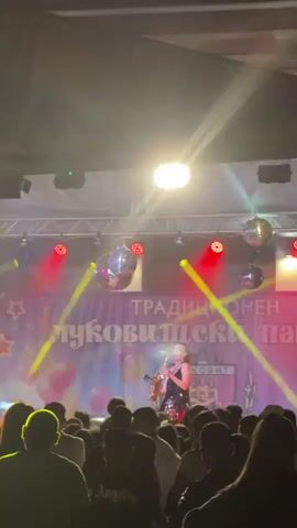 A concert is taking place. In the foreground, the backs of a crowd of people are visible. In the middle ground, a person on stage is playing a saxophone and singing into a microphone. They are wearing a dark, sequined outfit. Behind the singer, a banner with Cyrillic writing is displayed. The stage is decorated with disco balls and illuminated by red and yellow spotlights. A second person is visible on stage, to the left of the singer, wearing a white t-shirt and a hat, and appears to be dancing. The environment is dimly lit, characteristic of a performance venue.