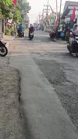 The video shows a street scene with several people on motorcycles. The road surface is uneven and appears to be under repair in some sections. Flags, likely national flags, are visible along the street, suggesting a festive or celebratory occasion. In the foreground, a motorcycle is parked on the left side of the road. Further down the street, several other motorcycles are in motion, with riders moving in the same direction. Some individuals are standing near the side of the road, possibly observing or engaged in commerce. The overall environment appears to be a residential or commercial street in a community.