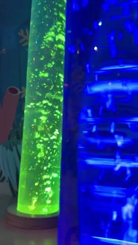 This media file features two tall, cylindrical bubble lamps that are illuminated and appear to be in operation. The lamp on the left is a vibrant green and filled with small, floating bubbles. The lamp on the right is a deep blue and shows a pattern of lights that move vertically, creating a cascading effect. The background is blurred, but suggests a colorful mural with elements resembling underwater life, such as coral and seaweed, painted in shades of blue and pink. The lighting is dim, characteristic of an indoor setting that might be designed for sensory exploration or relaxation. The overall mood is calming and mesmerizing due to the gentle movement of the bubbles and the vibrant, yet soft, colored lights. There is no visible text in the media file.