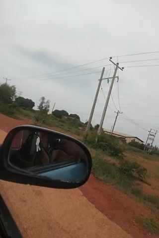 The video shows a side-view mirror of a vehicle traveling along a dirt road in Nigeria. The mirror reflects the interior of the vehicle, including car keys, a person's arm and torso wearing a brown shirt, and what appears to be a black dog. Through the mirror, the passing scenery is visible, characterized by lush green vegetation, trees, and a utility pole with wires. The road itself is a reddish-brown dirt track, indicating a rural or semi-rural setting. In the distance, more trees and some structures can be seen, possibly homes or other buildings. The sky is overcast with a pale, diffused light, suggesting it might be a cloudy day or late afternoon. The overall impression is of a casual drive through a landscape typical of the Nigerian countryside. There is no visible text in the media file.