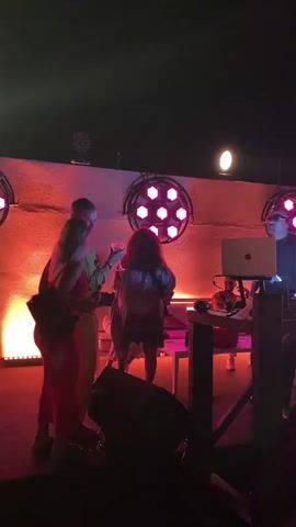 This is a short video clip of a live performance at night. The scene is illuminated by bright stage lights, predominantly in pink and white hues. In the foreground, to the left, a woman in a long orange dress stands with her back to the camera. To her right, a man in a light-colored shirt and pants is facing her. In the center, a person with dark, curly hair wears a patterned dress and appears to be dancing. Behind them, a white table is visible. To the right, a DJ setup is present with a laptop on a stand. The background features a textured wall with circular light fixtures. The overall atmosphere suggests a lively event, possibly a concert or party.