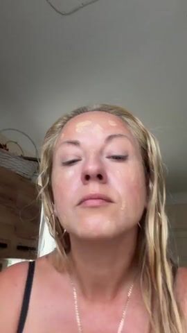 A woman is applying foundation to her face using a makeup sponge. She is looking down at her face as she blends the product. The woman has blonde hair and is wearing a black tank top and a necklace. The background appears to be a room with furniture.