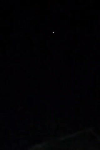 The video is a dark, night-time shot of the sky, with a single, bright, white light in the upper center of the frame. The light appears to be stationary. The bottom of the frame shows a dark, indistinct silhouette of what might be a roofline or building. The overall scene is dark and shows very little detail due to the low light conditions.