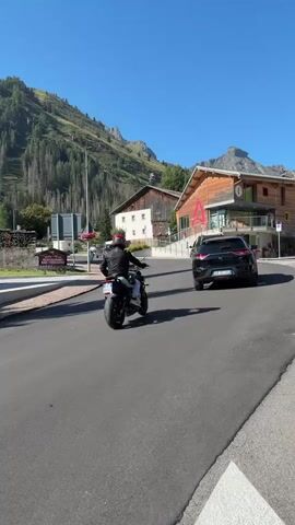 A motorcycle rider wearing a black helmet and a red jacket rides on a paved road. They are moving away from the camera and turning slightly to the right. The road is bordered by a sidewalk on the left. In the background, there are several buildings with wooden facades. To the left of the road, a signpost with multiple signs is visible. Behind the buildings, a large, green, forested mountain slopes upwards under a clear blue sky. There are a few cars parked and driving on the road. The sun is shining, casting shadows.