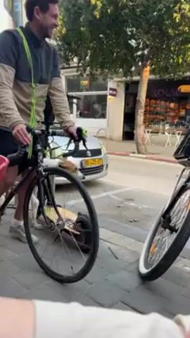 A video shows a man on a bicycle on the sidewalk. He is wearing a beige and gray fleece, black shorts, and white shoes. He has a yellow strap slung across his chest. The bicycle is black with a white frame. It is leaning against a bike rack. Another bicycle is parked beside it. A red plastic chair and a person are partially visible in the foreground on the left. Across the street, there is a shop front with the word “Kolbo” visible. Cars are parked along the street. The day appears sunny.