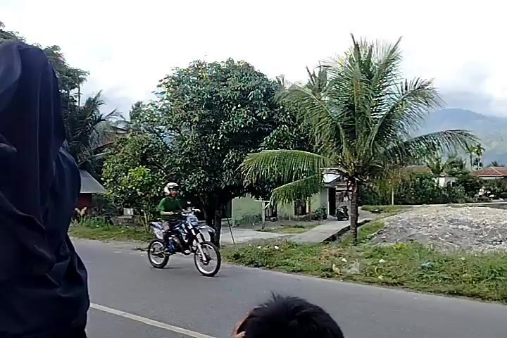 The video shows a street view from a moving vehicle. A person wearing a black hijab is in the foreground on the left, with a red and white garment partially visible on the far left. In the background, there are several small buildings with corrugated metal roofs, some vegetation, and palm trees. The road appears to be asphalt, with a white line marking the lane. The sky is overcast. Another person is partially visible in the lower right corner of the frame. The video is filmed at a low angle and appears to be handheld, suggesting it was recorded by a passenger in the vehicle. The scene suggests a rural or semi-urban environment.