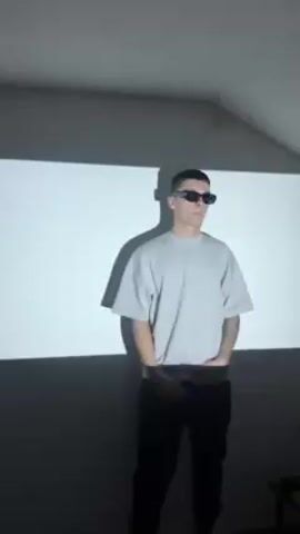 A young man wearing sunglasses, a gray oversized t-shirt, and black pants is dancing in a room. The background consists of a white wall onto which words are being projected. The words appear to be in a Slavic language.  The man makes motions with his arms and hips. A shadow of the man is visible on the wall, cast from the light source.