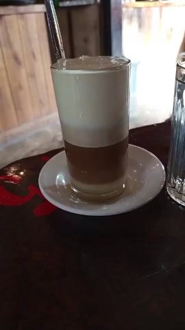 The video shows a glass of layered drink sitting on a white saucer on a wooden table. The drink has three distinct layers: a creamy white layer on top, a brown layer in the middle, and a clear liquid layer at the bottom. A metal spoon is stuck in the top layer of the drink. To the right of the glass is a clear, empty glass with a ridged design. The table appears to be dark wood with some red accents visible. In the background, there is a wooden structure resembling a bar or counter. The lighting is ambient, suggesting an indoor setting. The camera is static and focused on the drink.
