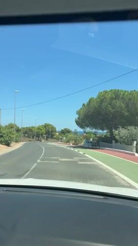 This media file captures a view from the inside of a vehicle, likely a car, driving along a road on a bright, sunny day. The scene is set in Alcalà de Xivert, Spain. The road is paved and has lane markings, with a bike lane to the right, distinguished by its green and red coloring. Several streetlights line the left side of the road.

The primary subjects are the road and its surroundings. In the distance, there are trees and vegetation, suggesting a semi-rural or coastal environment. The sky is a clear, vibrant blue, with a single contrail visible.

The activity is driving. The vehicle is moving forward on the road. There are other vehicles visible: a white truck, a red car, and a white car parked on the side of the road. There are also what appear to be speed bumps on the road surface.

Notable details include the clear blue sky and bright sunshine, indicating pleasant weather. The presence of a bike lane and streetlights suggests this is a developed area, possibly a town or a well-maintained route. The ocean is faintly visible in the far distance, hinting at a coastal proximity. There are no people clearly visible within the frame. The time of day appears to be midday due to the strong sunlight.

No visible text is present in the media file.