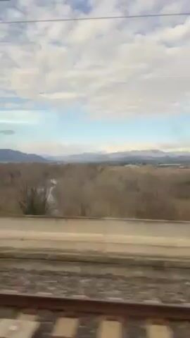 The video shows the view from a moving train window, passing by rural scenery. In the distance, there are mountains with snow-capped peaks partially obscured by clouds. Closer to the train track, there is a field of trees with bare branches, suggesting it might be late autumn or winter. The sky above is a mix of blue and white clouds. The railroad tracks are visible at the bottom of the frame, and a blurred barrier-like structure is seen in the foreground. The camera moves along with the train.