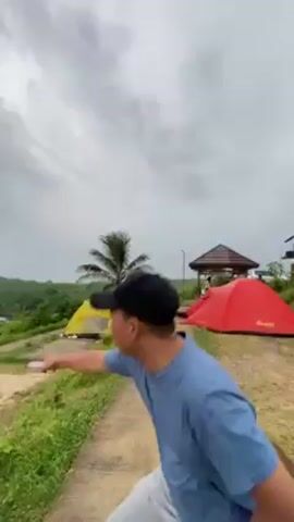 The video shows a man in a blue shirt and black cap, pointing towards the ocean from a campsite. There are three tents visible: red, grey, and yellow. The campsite is grassy with a paved walkway, with the tents erected on concrete pads.  A small gazebo stands near the red tent. A palm tree is located near the yellow tent. The sky is overcast. The camera pans to show the ocean view.