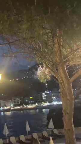 The video captures a nighttime scene with fireworks exploding in the sky over a coastal town. In the foreground, a pool area is visible with lounge chairs and umbrellas. A tree branch with pine needles partially obscures the view. The town below is lit up with lights, and the water reflects the lights from the town and the fireworks. The fireworks are white and pink bursts of light against the dark sky. A few people are visible in the lower right corner of the frame, seated near the pool. The overall atmosphere is festive and celebratory.