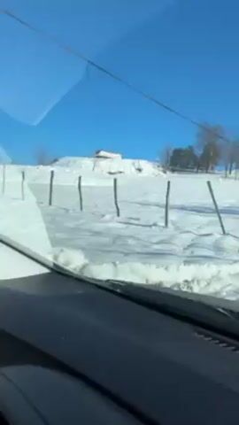A car is driving on a snow-covered road on a bright, sunny day. The road is narrow and lined with snowdrifts, some of which are taller than the hood of the car. The road slopes uphill.  Bare trees and utility poles are visible on either side of the road. In the distance is a snow-covered hillside with a small building at the top. The car's dashboard is visible in the foreground. Music plays softly in the background.