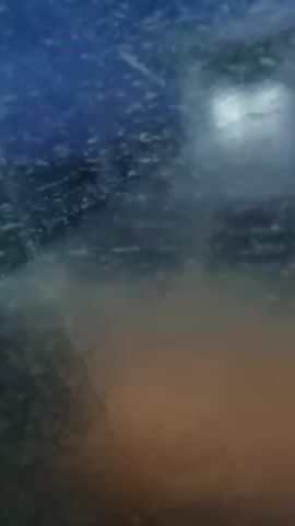 The video appears to be taken from inside a vehicle, looking out through a window covered in water droplets. The perspective is slightly angled upwards. Outside, it seems to be dark and possibly raining or snowing, obscuring the view.  A vehicle is visible through the water-streaked window. In the lower right corner, an amber or orange glow is present, possibly from a light source. A sticker is also visible on the bottom right of the window. The overall impression is one of limited visibility due to weather conditions.