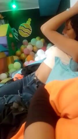 A medium shot shows a person in a white tank top and blue jeans sitting on a couch. The person has dark curly hair and is holding their arms up to their head. In the background, there are balloons and a play structure. The lighting is dim.