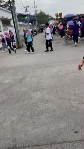 The video shows an outdoor scene with a group of people walking on a paved road. The people are diverse in age and gender, and many are wearing purple shirts. Some individuals are carrying bags or other items. In the background, there are buildings, market stalls, and power lines. The overall atmosphere appears to be a public gathering or event.