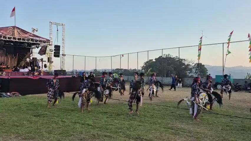 A group of people in traditional attire are performing a dance on a grassy field. They are wearing ornate costumes and some are holding props that resemble horses. The dance appears to be a cultural performance, possibly a traditional Javanese folk dance. In the background, there is a stage with musical equipment, a fence, and a crowd of spectators. The sky is clear and the sun appears to be setting. The overall scene suggests an outdoor festival or cultural event.