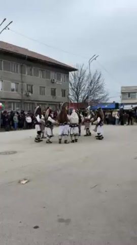 In an eye-level shot under an overcast sky, a circle of people dressed in traditional costumes perform in a town square. The costumes are predominantly white and brown, with tall headwear that appears to be made from animal fur. Each individual is holding what seems to be a percussion instrument, which they are using to create a rhythmic sound. A crowd of onlookers surrounds the performance, standing along the edges of the square. A multi-story building with visible air conditioning units is on one side of the square, while bare trees stand in the background. The ground is paved with concrete. The overall impression is of a cultural performance taking place in a public space.