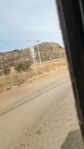 The video shows a ride along a road in what appears to be a three-wheeled vehicle such as a tuk-tuk or auto rickshaw. The road is paved but has some patches of dirt and what appears to be painted words on the surface. The landscape on either side of the road consists of dry grasses and low, rocky hills. There are power lines and utility poles along the road. The sky is overcast. The vehicle appears to be moving at a moderate speed.