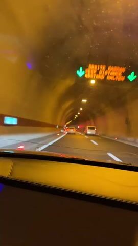 The video shows the interior of a car driving through a tunnel. The tunnel is lit with yellow overhead lights and has a smooth, curved ceiling. On the road ahead, other vehicles are visible, including cars and a white van. Red and white lane markers are present on the road. The dashboard of the car is visible in the foreground, with a quilted pattern and a yellowish glow. The overall impression is of movement through a man-made underground passage.