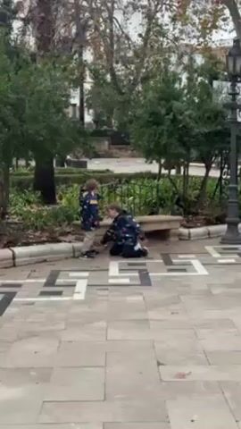 Two young children are in a park on a cloudy day. One child stands while the other kneels on the ground next to a stone bench. Both children wear dark jackets with a colorful pattern. The park has trees, grass, and ornamental plants. The ground is paved with stone tiles in a geometric pattern. A black metal lamppost stands to the right. One child speaks while the other listens.