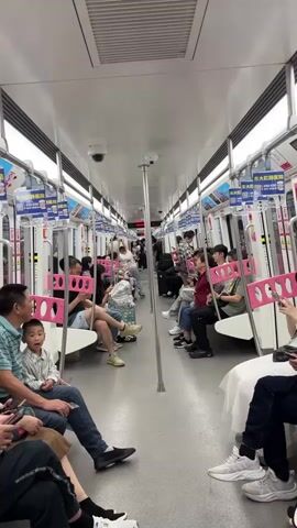 The interior of a subway car is shown in a stationary video. People are sitting and standing throughout the car. The seats are positioned facing each other along both sides of the subway car. Many of the passengers are using their phones. An advertisement is visible on the handrails above the seats. The interior of the subway car is white and gray with pink accents. A child is talking, and a subway bell can be heard.