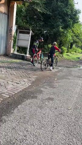 Two young individuals are engaged in an activity with bicycles in a semi-urban environment. One person, wearing a red and black outfit and a baseball cap, is standing next to a red and black bicycle, appearing to adjust it or show something on it to the other person. The second person, dressed in a red and black jacket and a beige cap, is seated on a black bicycle and looking towards the first individual. Both bicycles appear to be mountain bikes, suggesting a recreational or casual activity.

The scene is outdoors, with paved brick pathways and an asphalt road visible. Lush green trees and foliage dominate the background, indicating a day with good vegetation. The sunlight filtering through the trees and the general brightness suggest it is daytime, likely mid-morning or afternoon. The presence of puddles on the asphalt might indicate recent rain, but the overall lighting suggests it's not actively raining.

A light-colored structure, possibly a small shed or kiosk, is visible in the background, partially obscured by a tree. A metal gate or fence is also present to the left of the frame. Graffiti is visible on the gate, adding a touch of urban character to the scene. The ground is a mix of rough asphalt and patterned brickwork. The overall atmosphere appears relaxed and casual, with no indication of distress or urgency.

There is no discernible text in the visible portion of the media file. The setting appears to be a residential or recreational area in Siring, Indonesia, characterized by its blend of paved surfaces, natural greenery, and modest structures.