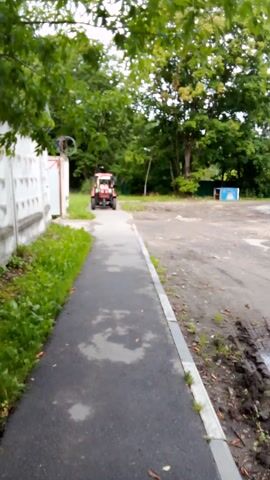 This media file shows a small, red and white tractor driving down a paved path. The tractor is the primary subject. It appears to be a utilitarian vehicle, possibly used for landscaping or light agricultural work. The scene is outdoors, with lush green trees and foliage lining the left side of the path. To the right of the path is a grey concrete border and a dirt or gravel area. A low white wall with a decorative pattern is visible along the far left edge of the frame, with some wire fencing and debris behind it.

The tractor is moving away from the camera and towards the left side of the image. The movement of the tractor is the main activity depicted.

The time of day appears to be daytime, likely late afternoon or early evening, given the soft lighting and the shadows cast. The ground looks slightly wet, suggesting it may have recently rained or there was dew. The location is identified as Arkhangel'skoye, Russia. There is no visible text in the media file. The overall mood is neutral, focusing on the mechanical operation of the tractor within its environment.