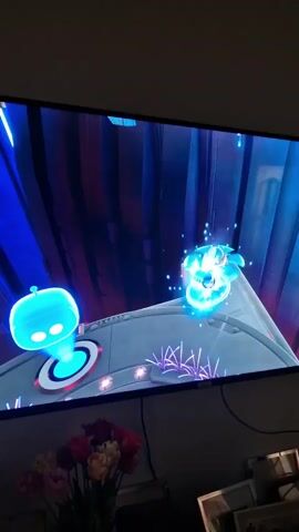 This is a video recording of a television screen displaying a video game. The game appears to be a 3D platformer with a futuristic or sci-fi aesthetic. The primary subject is a small blue robot character with glowing white eyes, possibly the player character. It is standing on a gray platform with circular glowing accents and some pink, spiky foliage. In the background, there are tall, abstract structures bathed in vibrant blue and purple light, suggesting an artificial or digital environment. A second, smaller robot with a similar glowing appearance is visible on another platform to the right.

The gameplay seems to involve navigating these platforms, possibly collecting items or reaching goals. One of the platforms has a holographic projection of a blue sphere with a white circle inside, and another has a transparent box with a glowing blue square inside. In the distance, a suspended platform with a green, grid-like structure holding some yellow spheres can be seen, indicating verticality and varied terrain. The overall mood of the game is bright and energetic, with a focus on vibrant colors and clean, futuristic design.

The video is shot from a slight angle, capturing the television screen and a portion of the room it is in. In the foreground, partially obscuring the bottom of the screen, is a bouquet of tulips in shades of pink, yellow, and red. The lighting in the room appears to be dim, with the primary illumination coming from the television screen itself. There is no discernible time of day or weather mentioned in the visual content. No people are directly visible within the game's scene, but the presence of the television suggests someone is playing it. No explicit text is visible within the game's interface or environment in these frames.