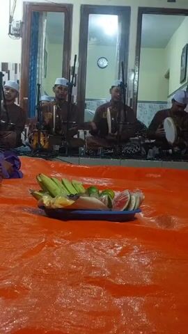 The video displays an indoor scene with four young men in matching brown robes and white caps seated in front of a wall with windows. They appear to be playing musical instruments, possibly rebana, and are positioned behind microphones. In the foreground, a blue platter holds a selection of cut fruit, including bananas, watermelon, and other unidentifiable items. The platter rests on an orange tarp spread across the floor. The overall impression is that of a musical performance or religious gathering.